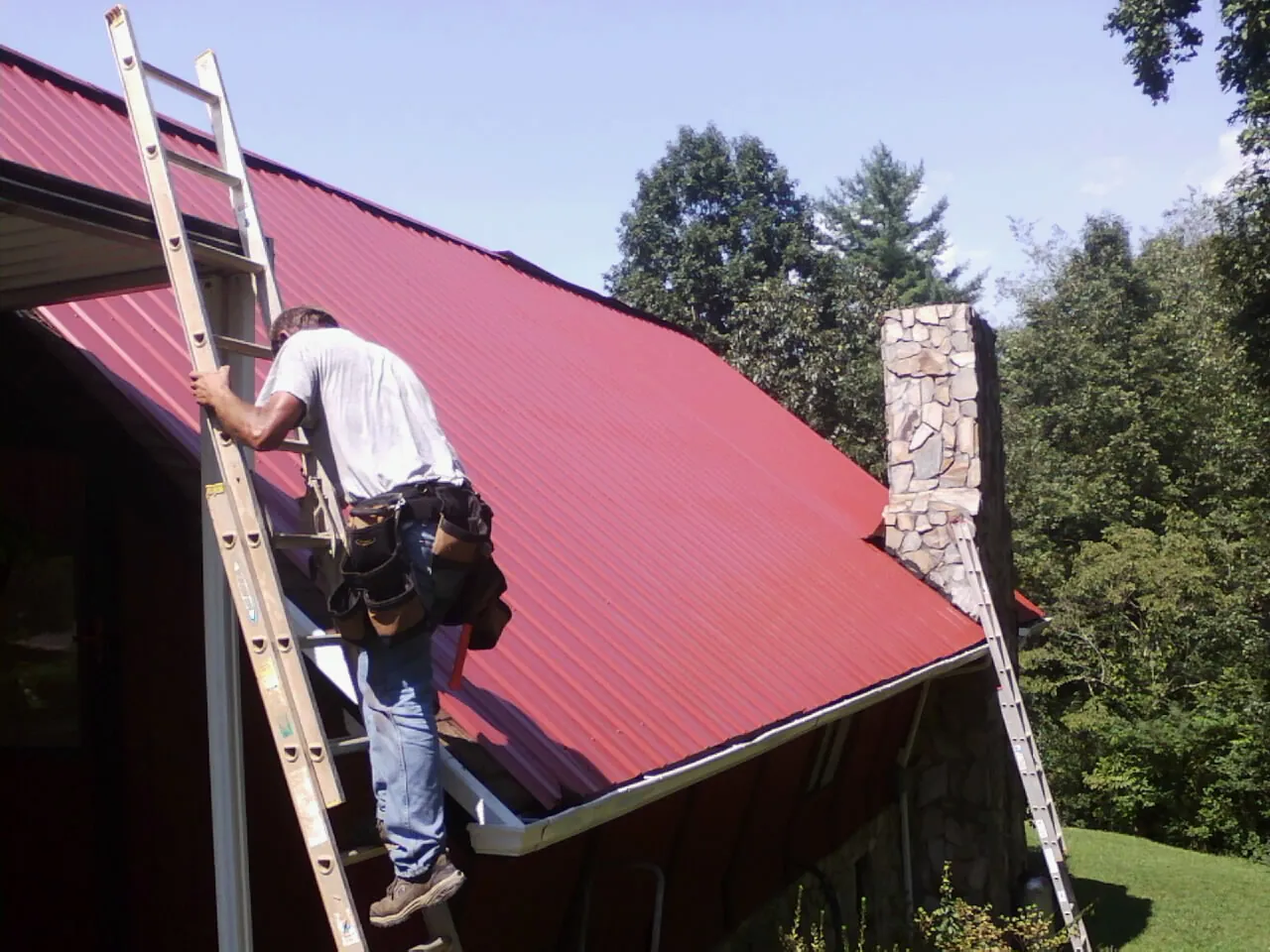 Roofer on a ladder beside a red metal roof and stone chimney — Soffit & Fascia Repair by Roofing Brothers in Muscoda